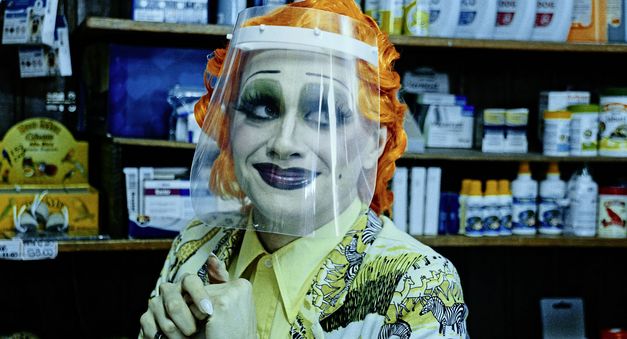 Filmstill aus „Three Tidy Tigers Tied a Tie Tighter“ von Gustavo Vinagre. Vor einem Ladenregal sitzt eine Person mit knalligem, künstlichem, orangenen Haar und lächelt zur Seite. Ihr Gesicht ist stark geschminkt und erinnert an einen Clown oder Drag-Shows. Vor dem Gesicht hängt ein Plastik-Schutzschild.