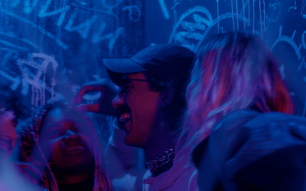 Film still from Sarnt Utamachote’s film “I Don’t Want to Be Just a Memory”. Five people laugh and move together in a room lit by blue and purple light, and a graffiti wall fills the background behind them. 