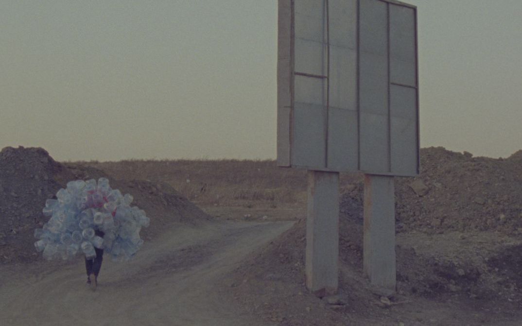 Film still from Hicham Gardaf’s film “In Praise of Slowness”. A person walks through a dry, barren landscape holding a mountain of empty plastic bottles on their back. To the right of the frame is what seems to be a large signpost, however we only see it from behind.