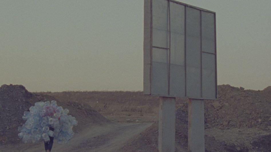 Film still from Hicham Gardaf’s film “In Praise of Slowness”. A person walks through a dry, barren landscape holding a mountain of empty plastic bottles on their back. To the right of the frame is what seems to be a large signpost, however we only see it from behind.
