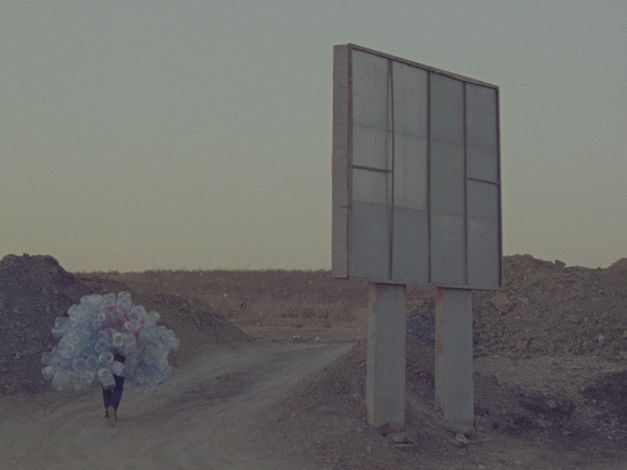 Film still from Hicham Gardaf’s film “In Praise of Slowness”. A person walks through a dry, barren landscape holding a mountain of empty plastic bottles on their back. To the right of the frame is what seems to be a large signpost, however we only see it from behind.