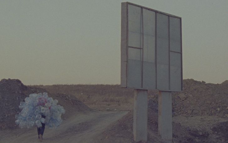 Film still from Hicham Gardaf’s film “In Praise of Slowness”. A person walks through a dry, barren landscape holding a mountain of empty plastic bottles on their back. To the right of the frame is what seems to be a large signpost, however we only see it from behind.