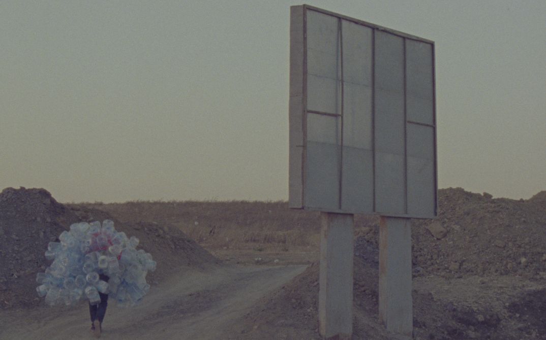 Film still from Hicham Gardaf’s film “In Praise of Slowness”. A person walks through a dry, barren landscape holding a mountain of empty plastic bottles on their back. To the right of the frame is what seems to be a large signpost, however we only see it from behind.