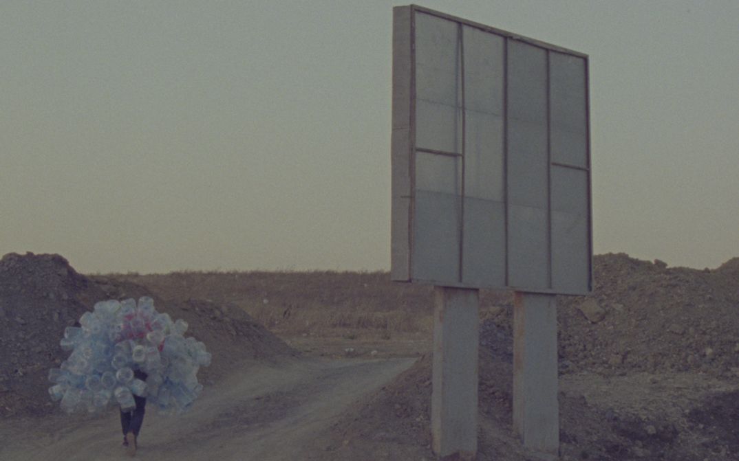 Film still from Hicham Gardaf’s film “In Praise of Slowness”. A person walks through a dry, barren landscape holding a mountain of empty plastic bottles on their back. To the right of the frame is what seems to be a large signpost, however we only see it from behind.