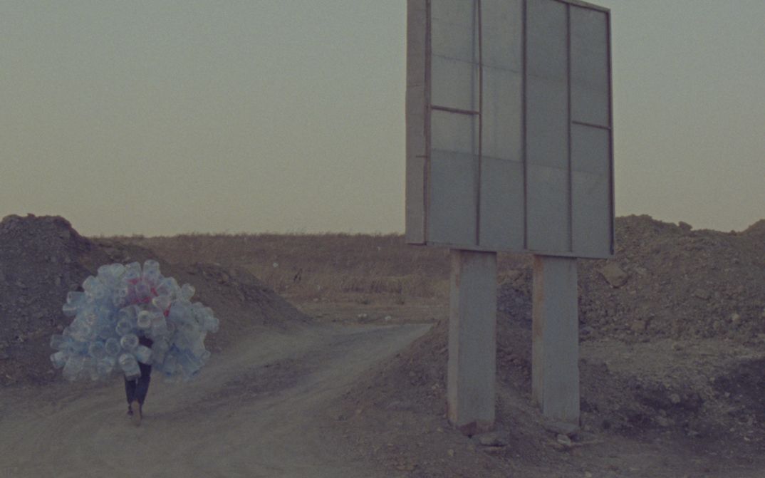 Film still from Hicham Gardaf’s film “In Praise of Slowness”. A person walks through a dry, barren landscape holding a mountain of empty plastic bottles on their back. To the right of the frame is what seems to be a large signpost, however we only see it from behind.