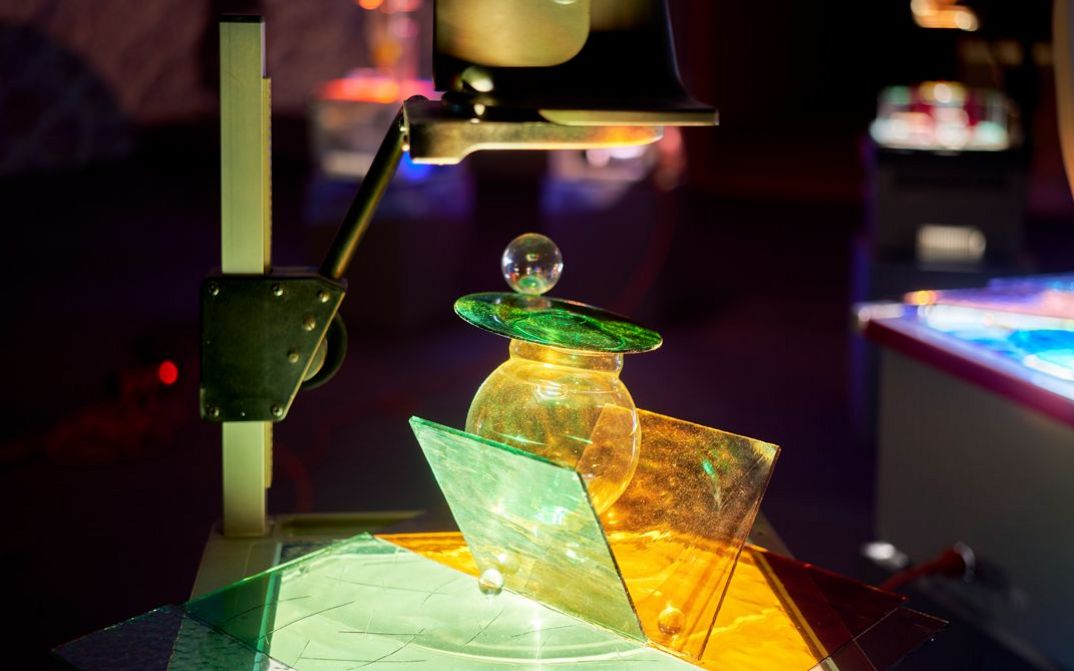 Filmstill aus dem Film „Sinking Suns“ von Neda Saeedi. Nahaufnahme einer Ausstellungsinstallation. Ein Projektor strahlt Licht aus, das von einer getönten Glasstruktur reflektiert wird.