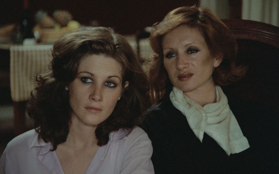 Filmstill aus SALE HISTOIRE: Zwei Frauen sitzen an einem Tisch und hören jemandem zu.