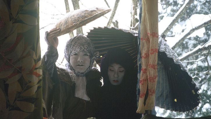 Film still from UNDER SNOW: Two people wearing makeup and costumes look out of a house window at a snowy landscape.