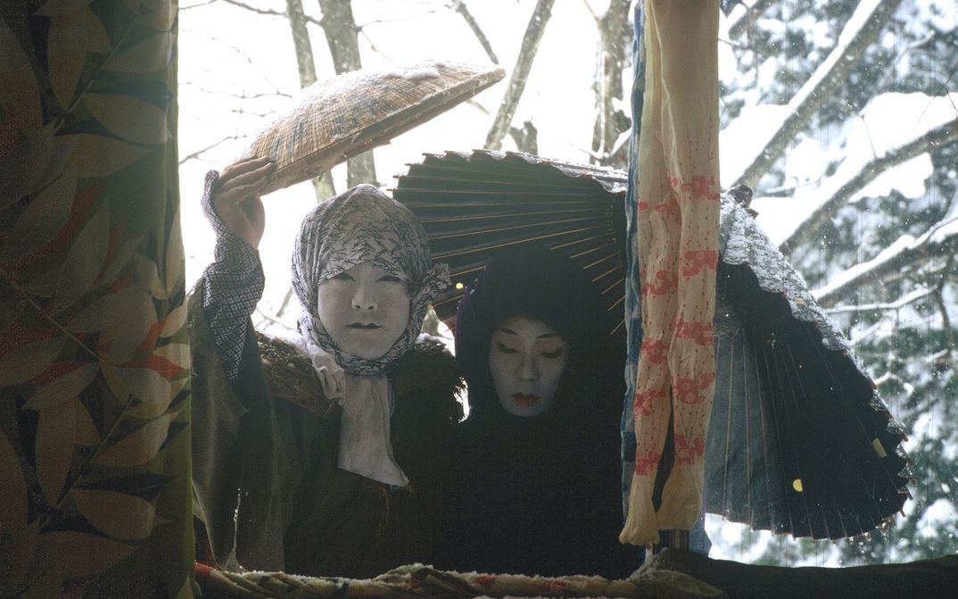 Film still from UNDER SNOW: Two people wearing makeup and costumes look out of a house window at a snowy landscape.