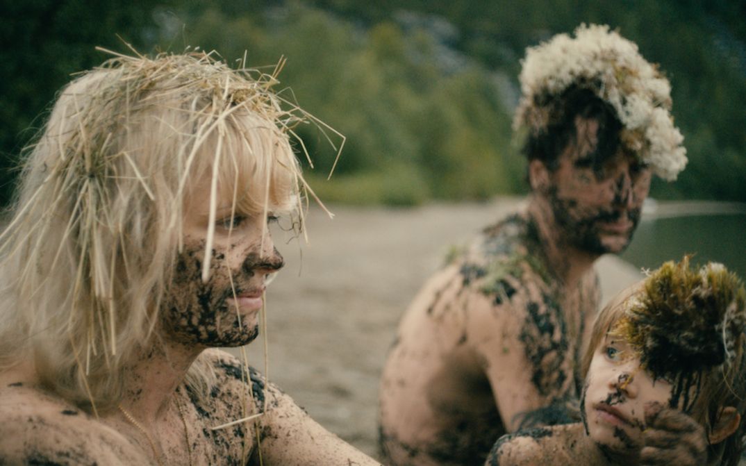Filmstill aus HEDI SCHNEIDER STECKT FEST: Eine Frau, ein Mann und ein Kind sitzen nackt an einem Ufer. Ihre Gesichter und Körper sind mit Schlamm beschmiert und in den Haaren steckt Stroh.