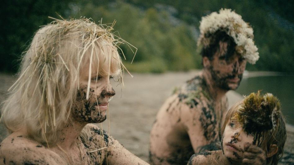 Filmstill aus HEDI SCHNEIDER STECKT FEST: Eine Frau, ein Mann und ein Kind sitzen nackt an einem Ufer. Ihre Gesichter und Körper sind mit Schlamm beschmiert und in den Haaren steckt Stroh.
