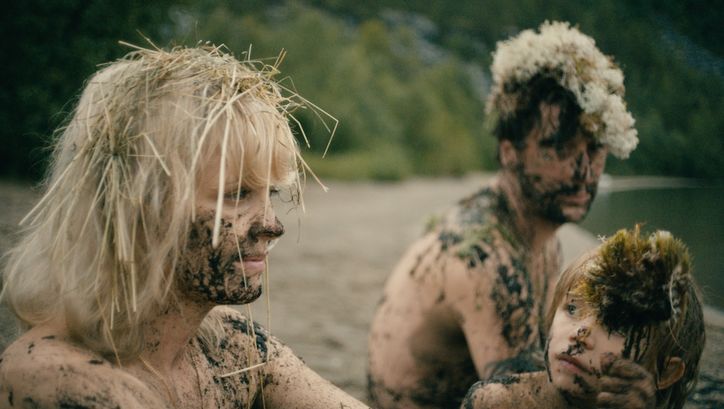 Filmstill aus HEDI SCHNEIDER STECKT FEST: Eine Frau, ein Mann und ein Kind sitzen nackt an einem Ufer. Ihre Gesichter und Körper sind mit Schlamm beschmiert und in den Haaren steckt Stroh.