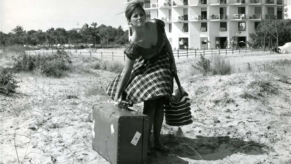 Film still from LA RAGAZZA CON LA VALIGIA: A young woman stands in a meadow with a large suitcase; a house can be seen in the background.