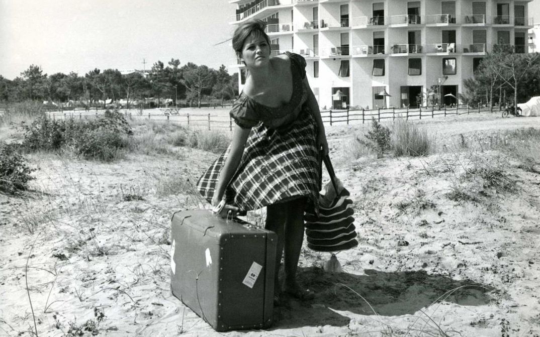 Film still from LA RAGAZZA CON LA VALIGIA: A young woman stands in a meadow with a large suitcase; a house can be seen in the background.