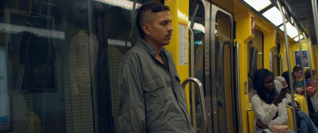 Mann steht in gelbem U-Bahn-Wagen, andere Fahrgäste sitzen im Hintergrund.