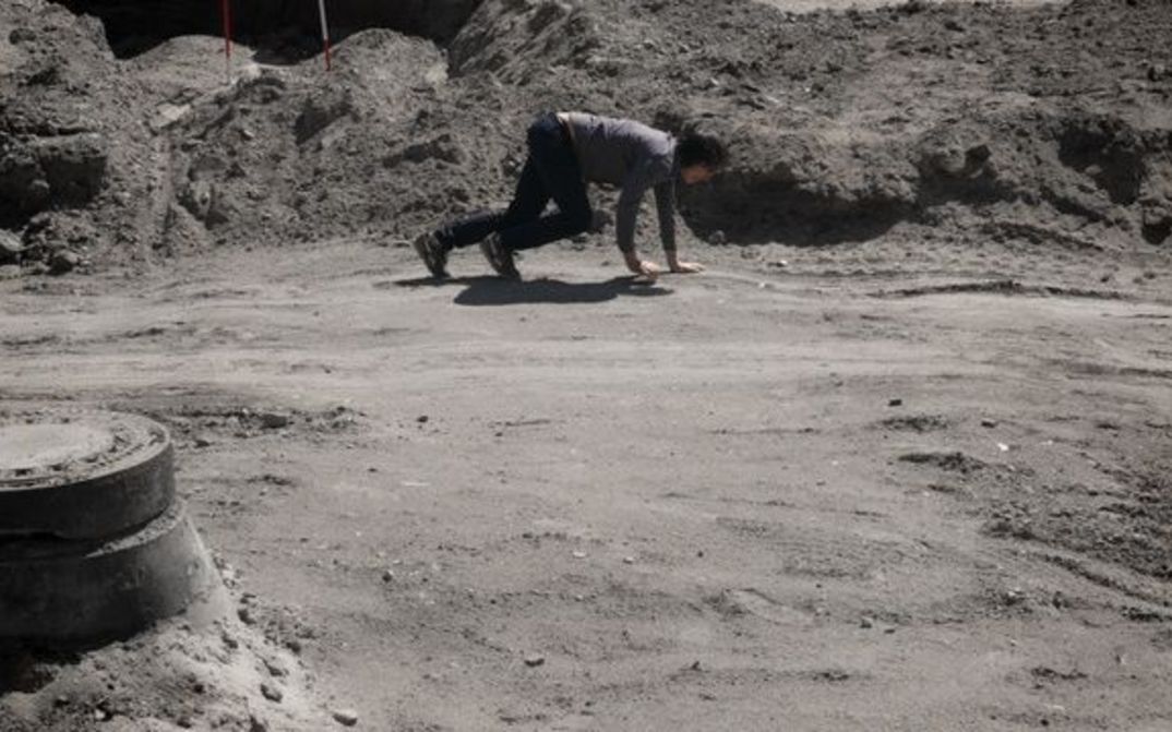 Film still from F ZWISCHEN DEN STUFEN DES ORGANISCHEN: A man on all fours between boulders.