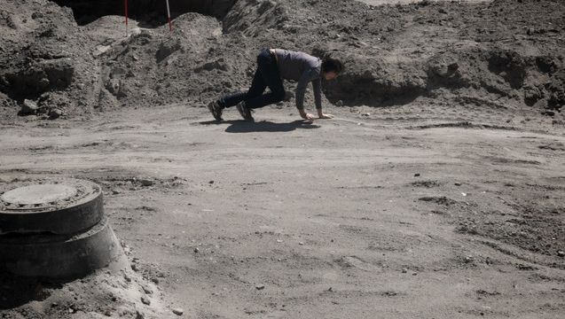 Film still from F ZWISCHEN DEN STUFEN DES ORGANISCHEN: A man on all fours between boulders.