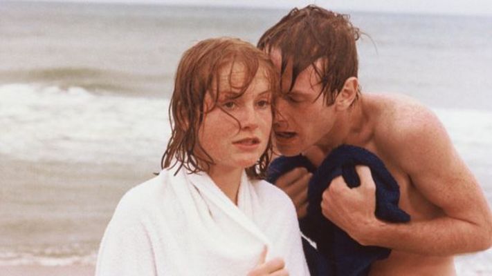 Filmstill aus LA DENTELLIÈRE: Ein junger Mann und eine junge Frau am Strand, er sagt ihr etwas ins Ohr.