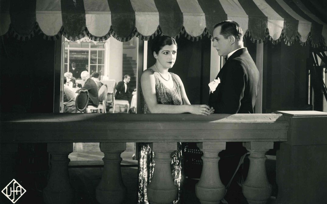 Film still from DIE DAME MIT DEM TIGERFELL: A man and a woman in elegant evening dress are standing on a terrace.