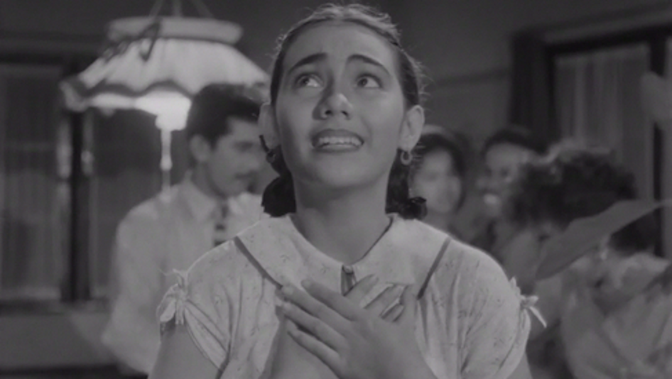 Filmstill aus TIGA DARA: Eine junge Frau hält sich die Hände vor die Brust und blickt nach oben.