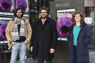 Three people are standing outside in front of a building with a large window. In the window, we can see the same poster several times. It is purple, there is a dragon fruit.