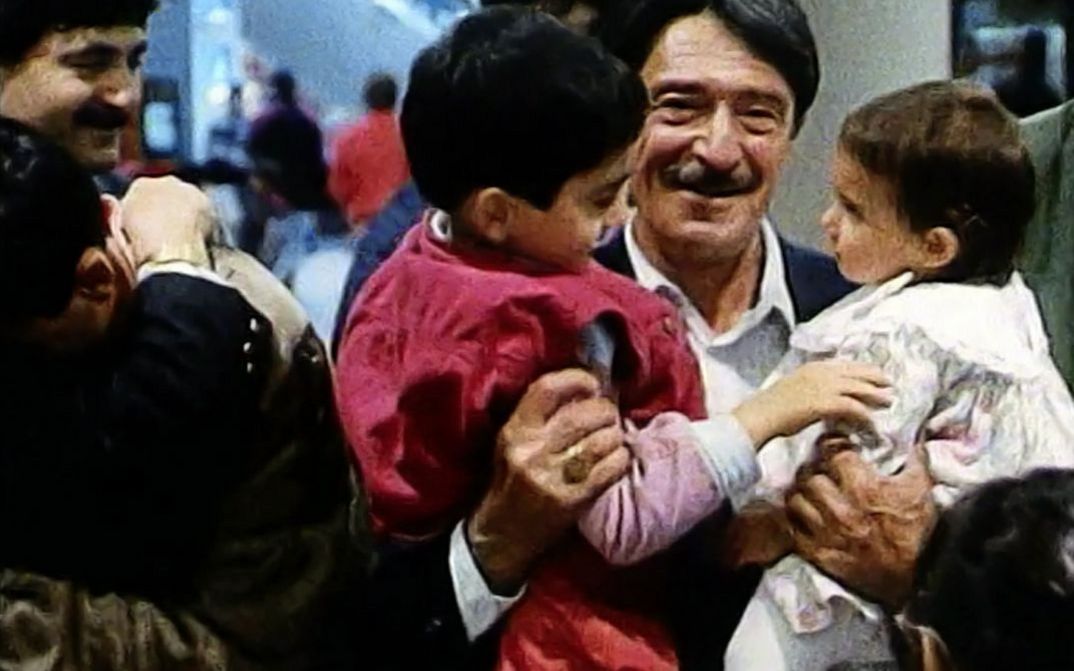 Still from the film „Mein Vater, der Gastarbeiter“ by Yüksel Yavuz. A man with a moustache holds two children in his arms and smiles. On the left side of the picture, two people are hugging each other. Several people are standing in the background.