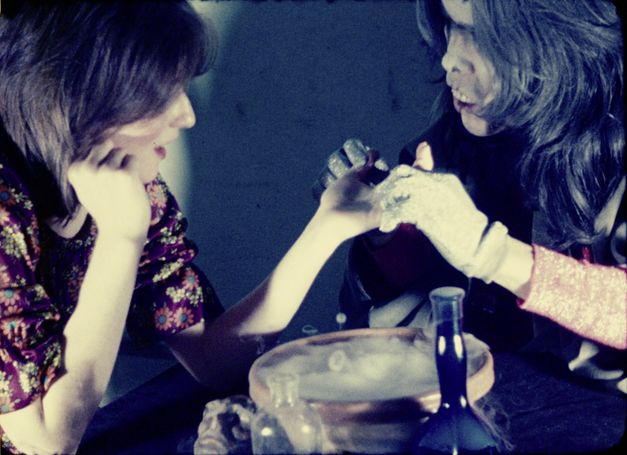 Filmstill aus "Palmistry" von Maria Lassnig. Zu sehen sind zwei Personen, die sich gegenübersitzen. Die Person auf der linken Seite trägt Glitzerhandschuhe und hält die Hand der Person auf der rechten Seite. 