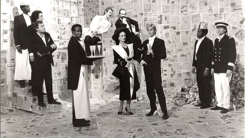 Filmstill aus DORIAN GRAY IM SPIEGEL DER BOULEVARDPRESSE: Eine Gruppe von elegant gekleideten Menschen und einige Kellner stehen in einem vollständig mit Zeitungen ausgekleideten Raum.