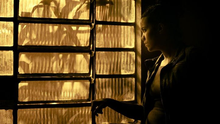A young woman stands at the side of a window made of fluted glass and looks out. The image is bathed in yellowish light. 