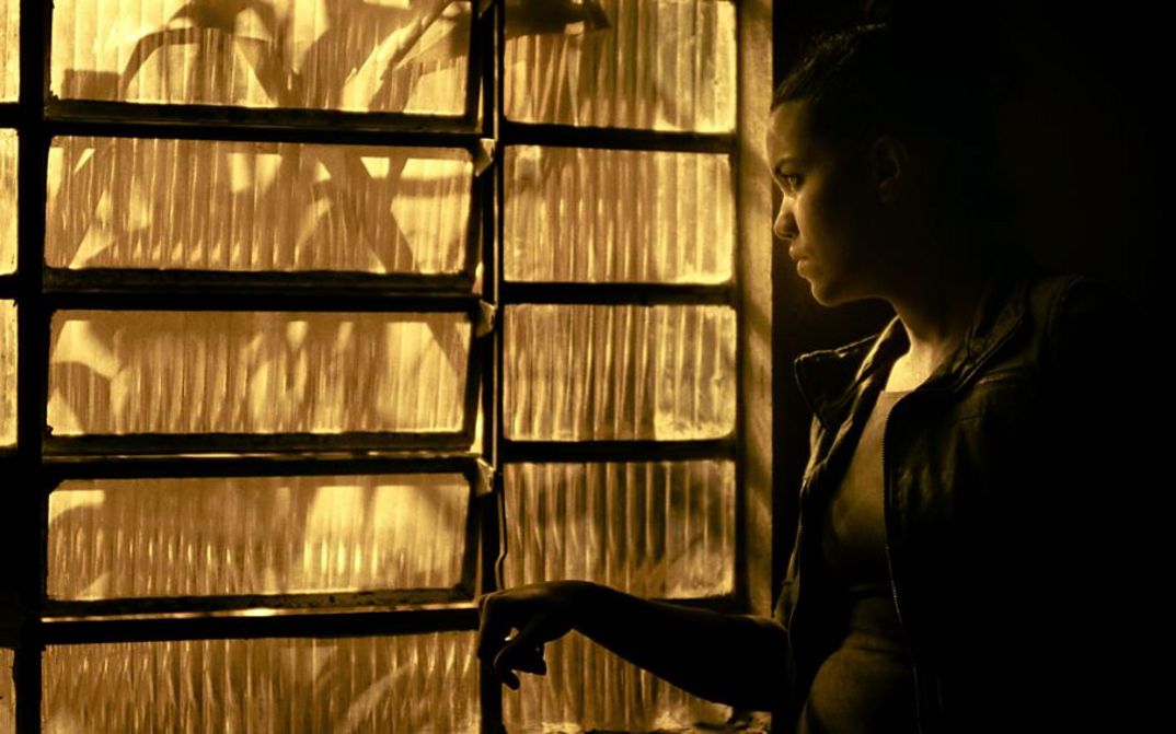 A young woman stands at the side of a window made of fluted glass and looks out. The image is bathed in yellowish light.