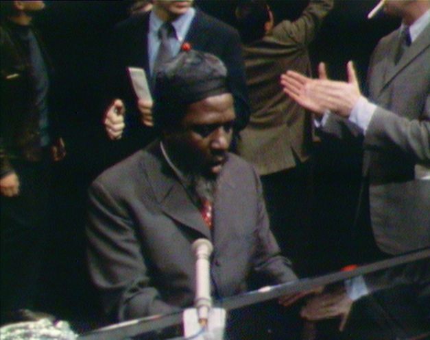 Still from the film “Rewind & Play“. A video image of Thelonious Monk playing the piano. Behind him we can see a few men in suits, one of them is clapping. On the piano, there is a microphone.