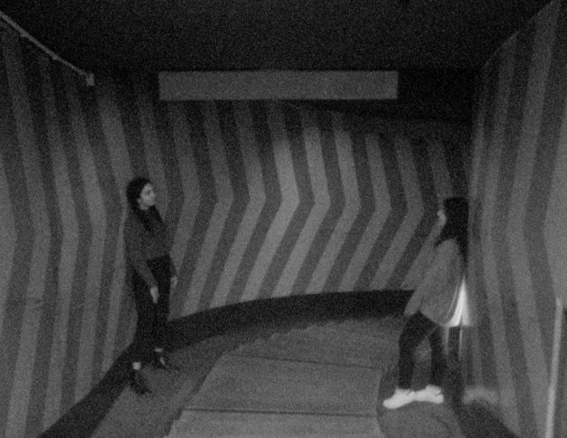 Filmstill aus „This Makes Me Want to Predict the Past“ von Cana Bilir-Meier. Ein Schwarz-Weiß-Bild von zwei Mädchen in einem Treppenhaus oder einer Unterführung. 