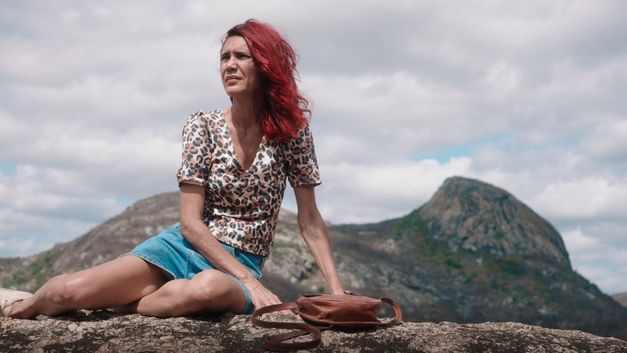 Frau mit roten Haaren sitzt auf einem Felsen in bergiger Landschaft, neben ihr liegt eine Tasche.