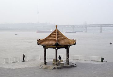 Filmstill aus Shengze Zhus „A River Runs, Turns, Erases, Replaces". Vor einem großen, grauen Fluss in diesigem Wetter steht eine Pagoda, von der aus zwei Menschen auf das Wasser gucken.