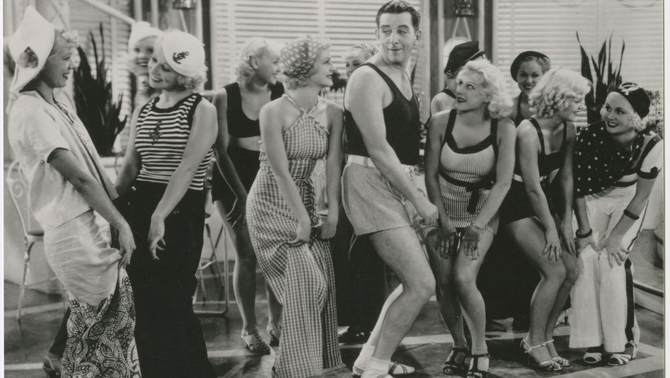 Filmstill aus THE GAY DIVORCEE: Edward Everett Horton ist in kurzen Hosen tanzend inmitten einer Gruppe von jungen Frauen.