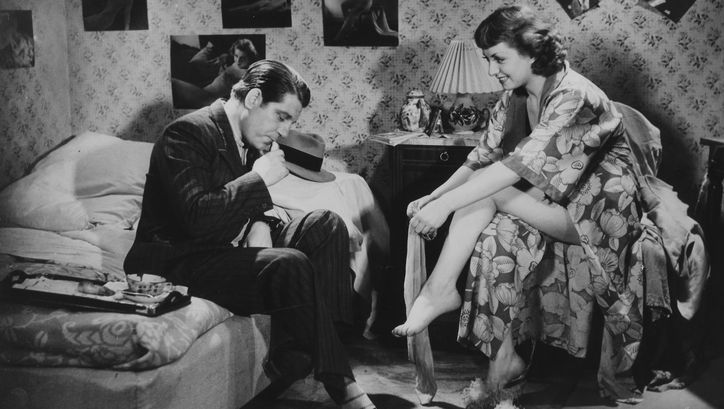 Film still from LA BELLE ÉQUIPE: A man and a woman in a bedroom. He sits on the bed in a suit, she sits opposite him in a bathrobe and puts on stockings.