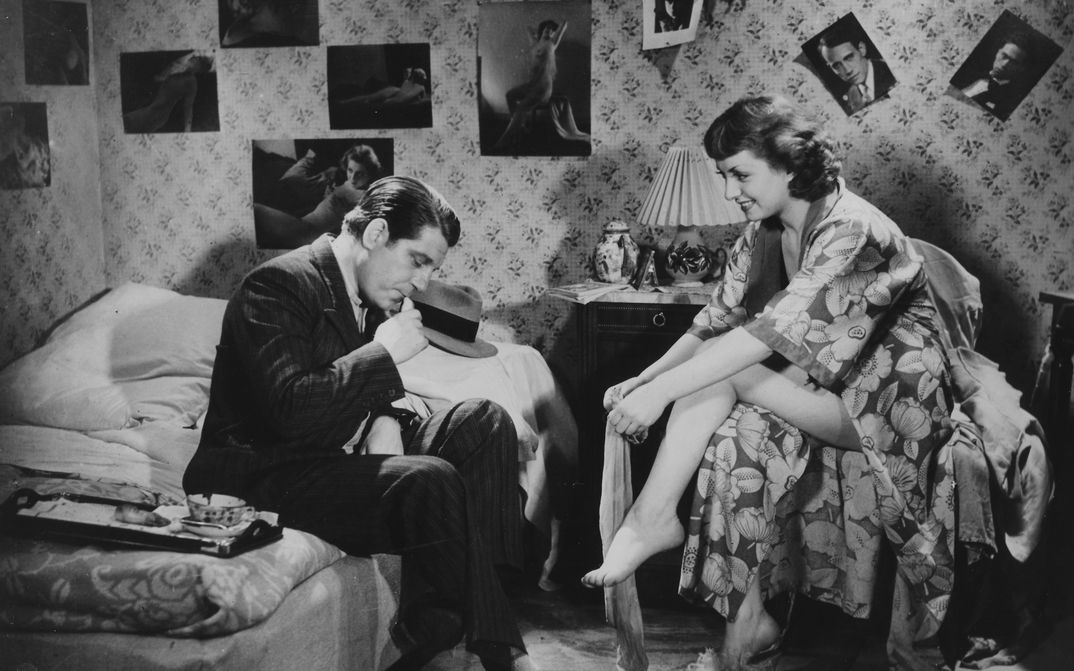 Film still from LA BELLE ÉQUIPE: A man and a woman in a bedroom. He sits on the bed in a suit, she sits opposite him in a bathrobe and puts on stockings.
