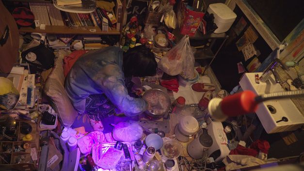Filmstill aus "Republic" von Jin Jiang. Zu sehen ist ein komplett überfüllter Raum aus der Vogelperspektive. In der Mitte sitzt eine Person und kocht. 