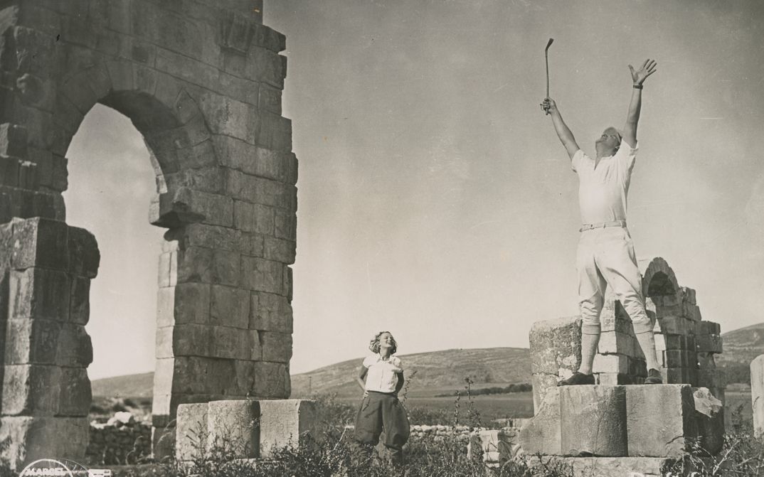 Filmstill aus DIE FÜNF VERFLUCHTEN GENTLEMEN: Ein Mann steht auf einer Ruine und streckt die Hände in die Höhe, eine Frau im Hintergrund schaut ihm zu.