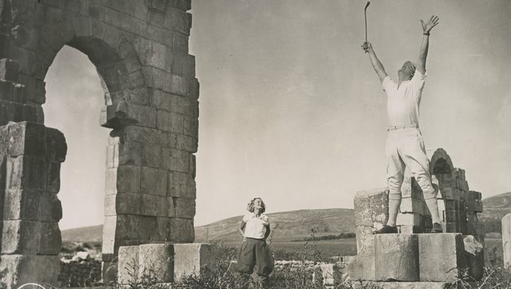Film still from DIE FÜNF VERFLUCHTEN GENTLEMEN: A man stands on a ruin and stretches his hands in the air, a woman in the background watches him.