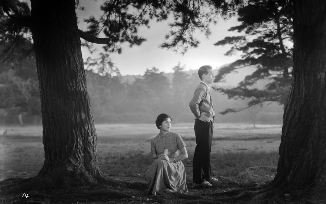 Filmstill aus TSUKI WA NOBORINU: Eine Frau und ein Mann auf einer Wiese zwischen zwei Bäumen, beide blicken rechts in die Ferne.