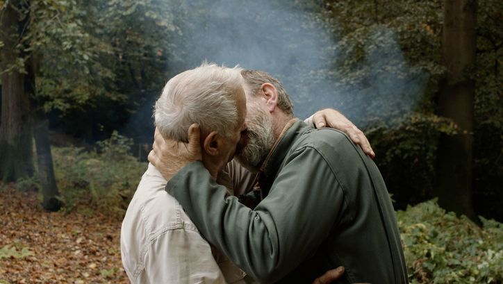 Film still from UN PRINCE: Two men kissing passionately. Forest and fog in the background.