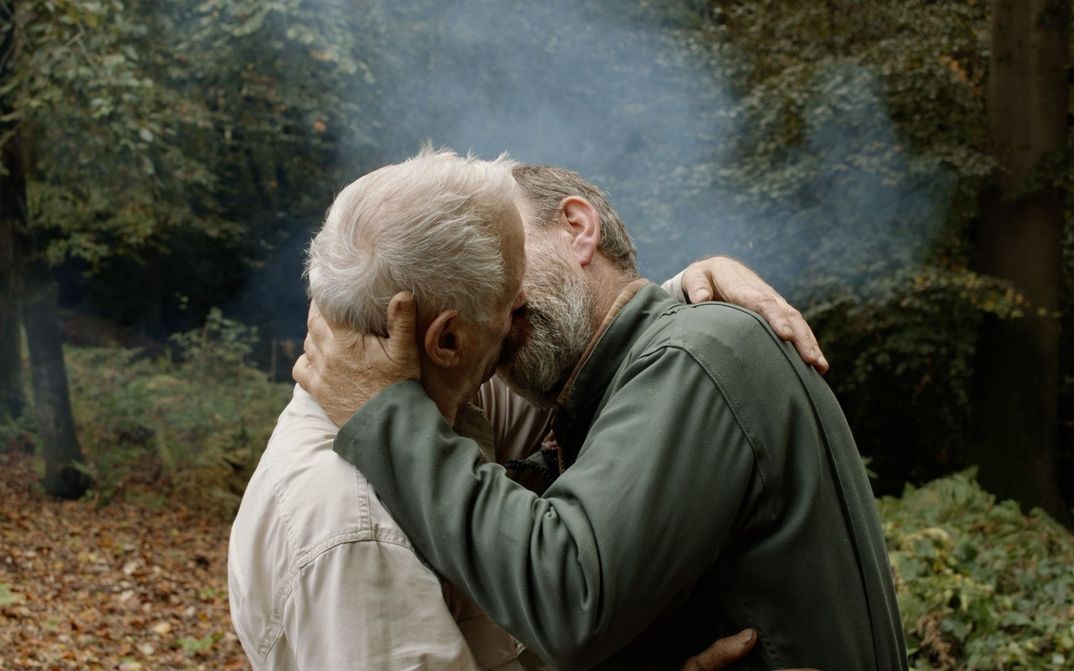 Film still from UN PRINCE: Two men kissing passionately. Forest and fog in the background.