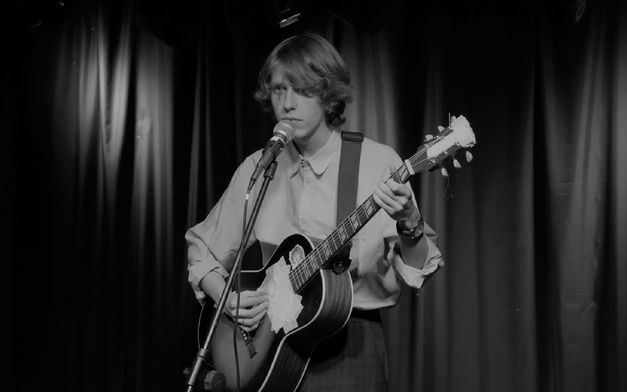 Film still from THE SWAN SONG OF FEDOR OZEROV by Yuri Semashko. The black and white image shows the protagonist at a microphone with a guitar.