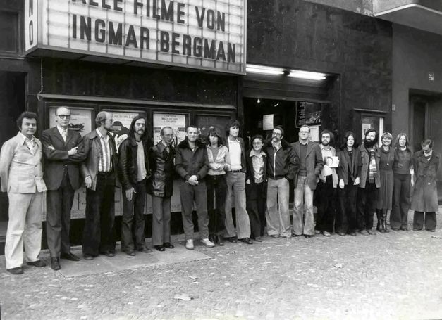 Filmstill aus „Komm mit mir in das Cinema – Die Gregors“ von Alice Agneskirchner. Ein Schwarz-Weiß-Bild einer Gruppe von Menschen nebeneinander aufgereiht vor einem Kino (dem Arsenal in der Welserstraße in Berlin). 