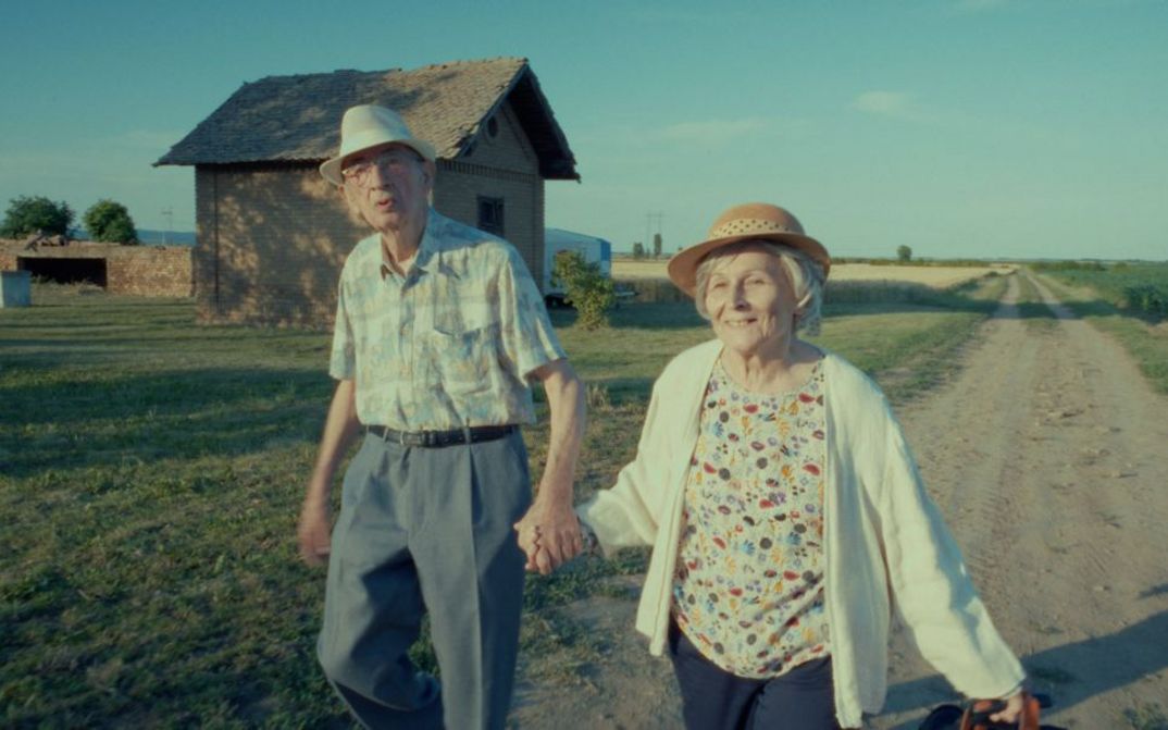 Filmstill aus RESTITUCIJA, ILI, SAN I JAVA STARE Gare von Želimir Žilnik. Das Bild zeiht eine ältere Frau und einen älteren Mann Hand in Hand auf einem unbefestigten Weg. Im Hintergrund ein einzeln stehendes Haus.