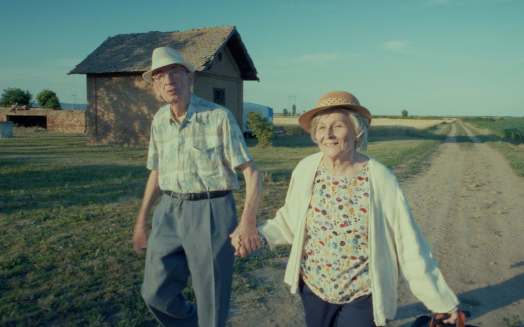 Film still from EIGHTY PLUS by Želimir Žilnik. The picture shows an elderly woman and an elderly man walking hand in hand along a dirt road. In the background is a single house.