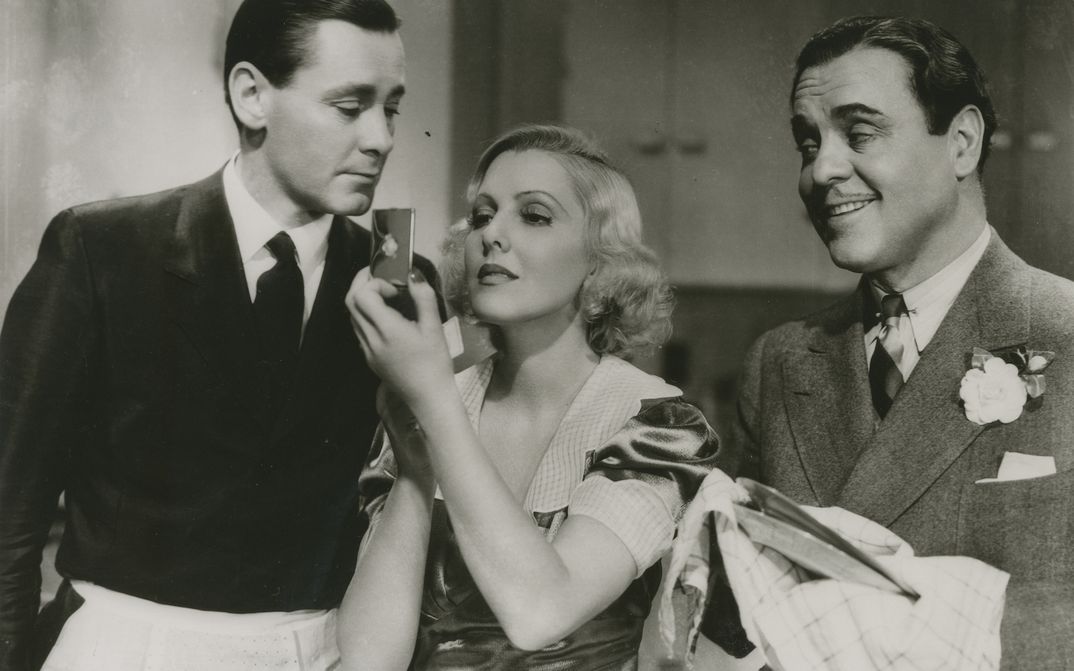 Film still from IF YOU COULD ONLY COOK: Two men and a woman are standing in a kitchen. The men are wearing aprons, one is drying dishes. The woman is looking in a hand mirror.