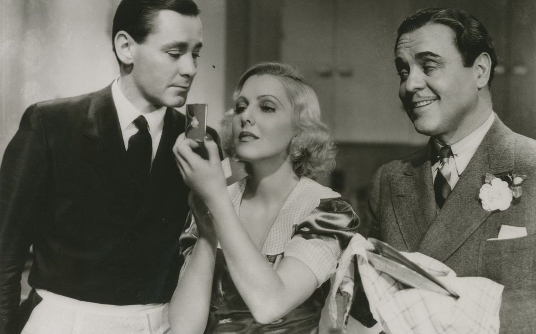Film still from IF YOU COULD ONLY COOK: Two men and a woman are standing in a kitchen. The men are wearing aprons, one is drying dishes. The woman is looking in a hand mirror.