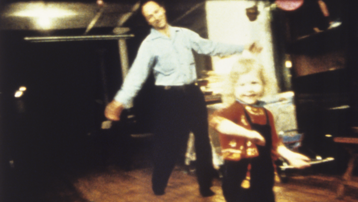 Film still from PARADISE NOT YET LOST: Jonas Mekas and a small child dancing in a room.